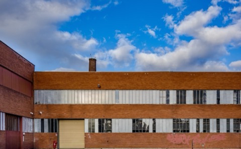 Façade d'un bâtiment industriel en brique rouge, deux niveaux de fenêtres en verre armé, une porte de garage jaune pâle au rez-de-chaussée, une cheminée de brique dépassant du toit, un tag rose sur le mur, ciel bleu chargé de nuages blancs.