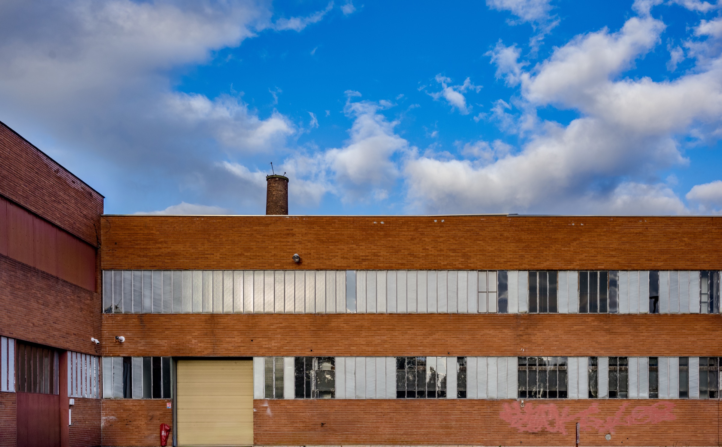 Façade d'un bâtiment industriel en brique rouge, deux niveaux de fenêtres en verre armé, une porte de garage jaune pâle au rez-de-chaussée, une cheminée de brique dépassant du toit, un tag rose sur le mur, ciel bleu chargé de nuages blancs.