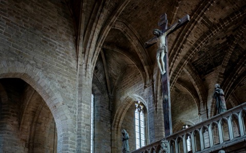 Un christ en croix sculpté, suspendu au sommet du jubé de pierre de l'abbatiale Saint-Robert de la Chaise-Dieu, sous des voûtes gothiques en pierre claire ; à droite, une statue en prière éclairée par un vitrail, dans la pénombre de la nef.