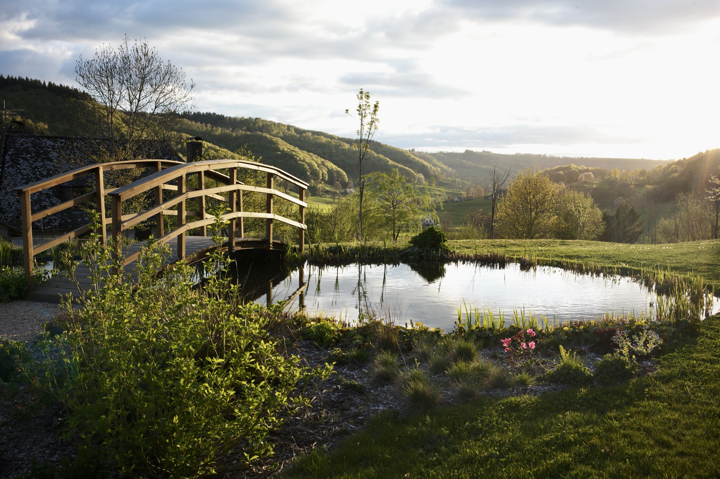 Un pont en bois arqué, à l'allure japonaise, enjambe un étang bordé de plantes et de fleurs roses, avec en arrière-plan une vallée verdoyante et vallonnée baignée d'une lumière rasante.