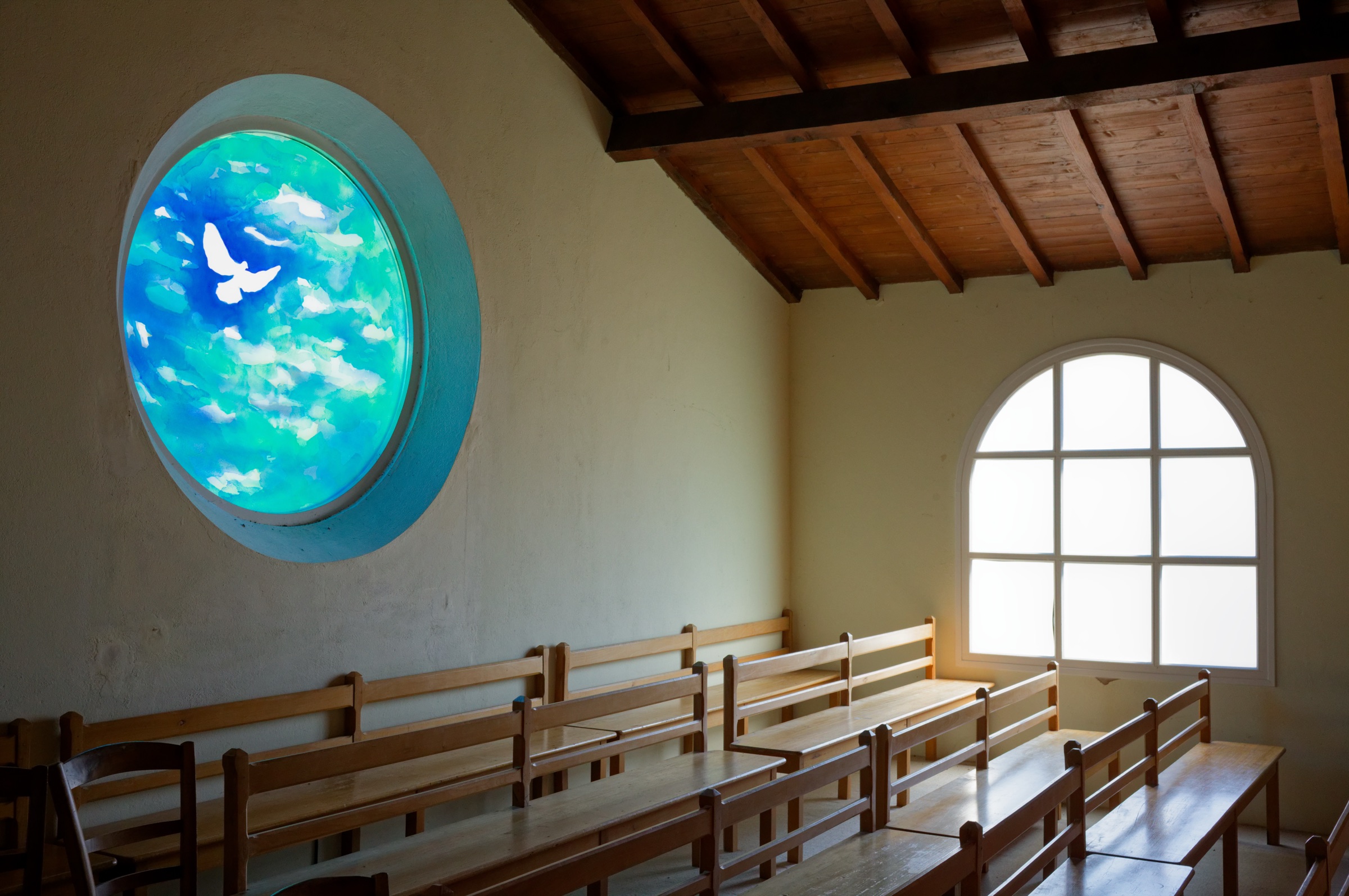 Intérieur de la chapelle Notre-Dame-du-Bois-de-la-Chaise, à Noirmoutier : bancs en bois vides sous une charpente apparente, un vitrail rond représentant une colombe blanche sur fond bleu et turquoise, une fenêtre en arc laissant entrer une lumière vive.